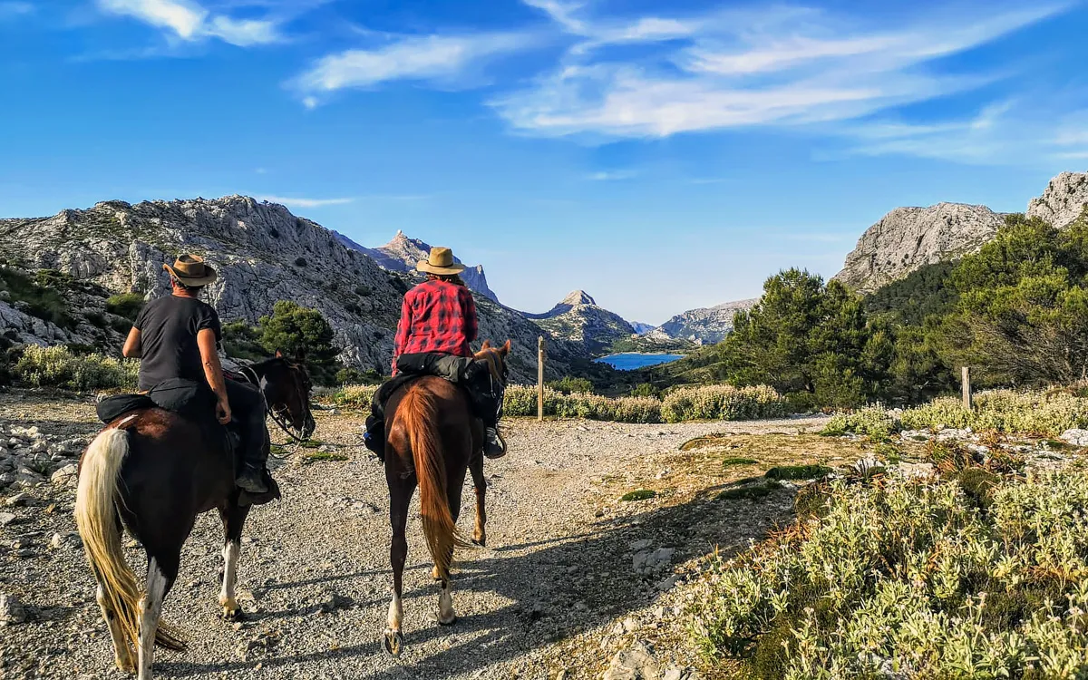 Deux cavaliers a cheval avancent dans un paysage montagneux et méditerranéen vers une crique d'eau bleue apercue au loin.