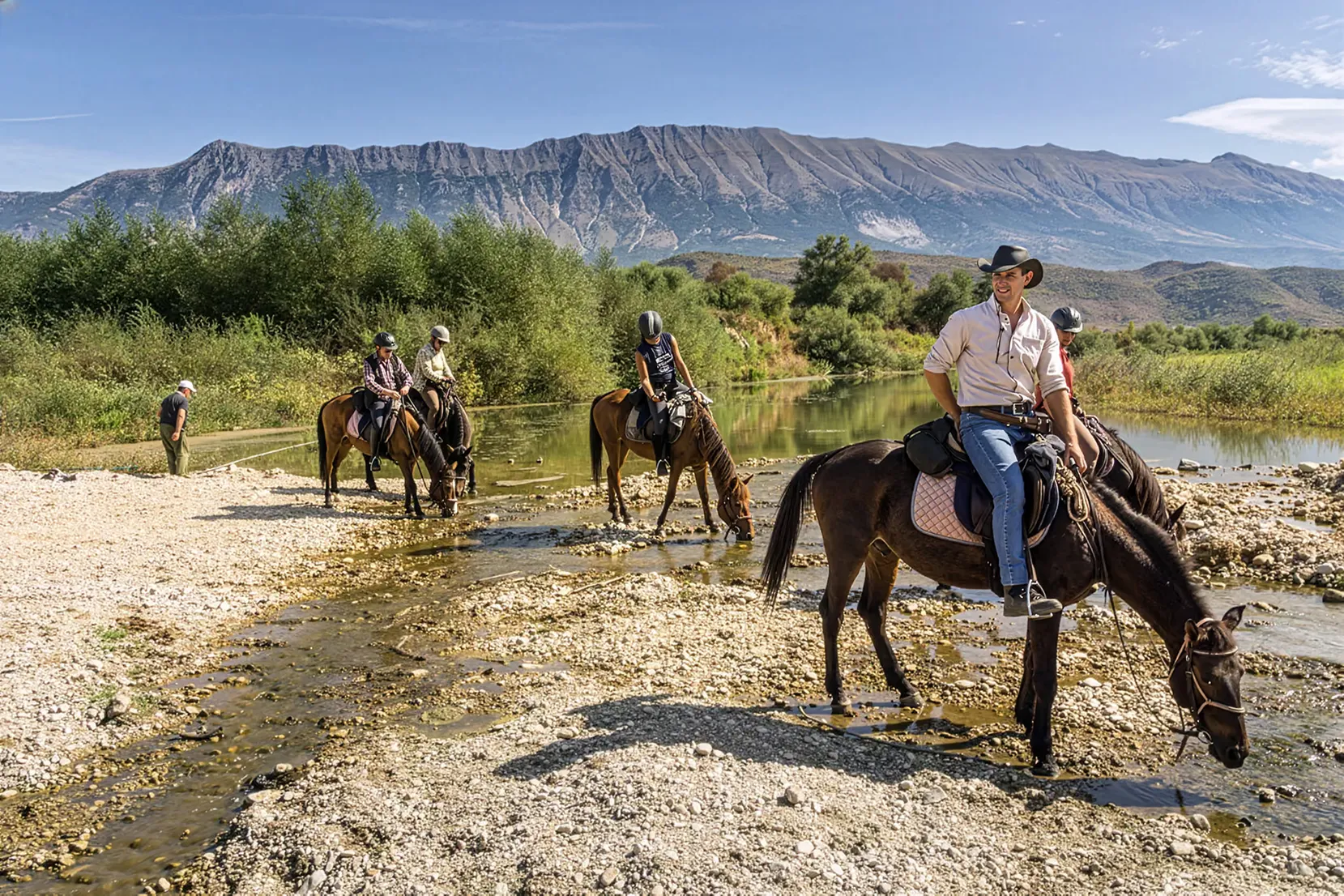 Guide équestre accompagnant des cavaliers à cheval lors d'une randonnée équestre en Albanie, traversant un ruisseau de montagne.