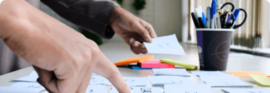 Personne organisant des notes manuscrites et des post-it colorés sur un bureau avec un récipient contenant des stylos et des ciseaux.