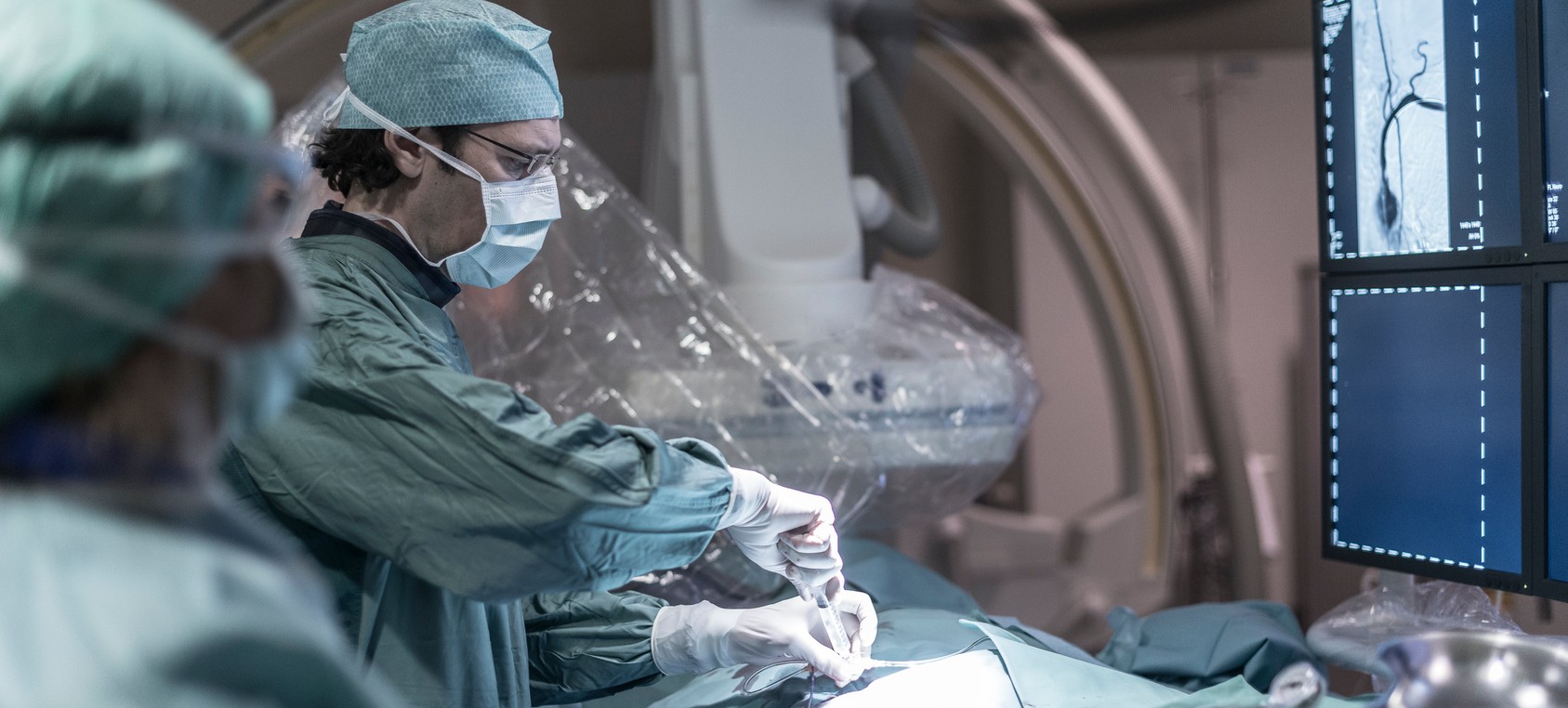 Surgeon in blue scrubs and mask handling a catheter delivery device over a draped patient during an interventional procedure