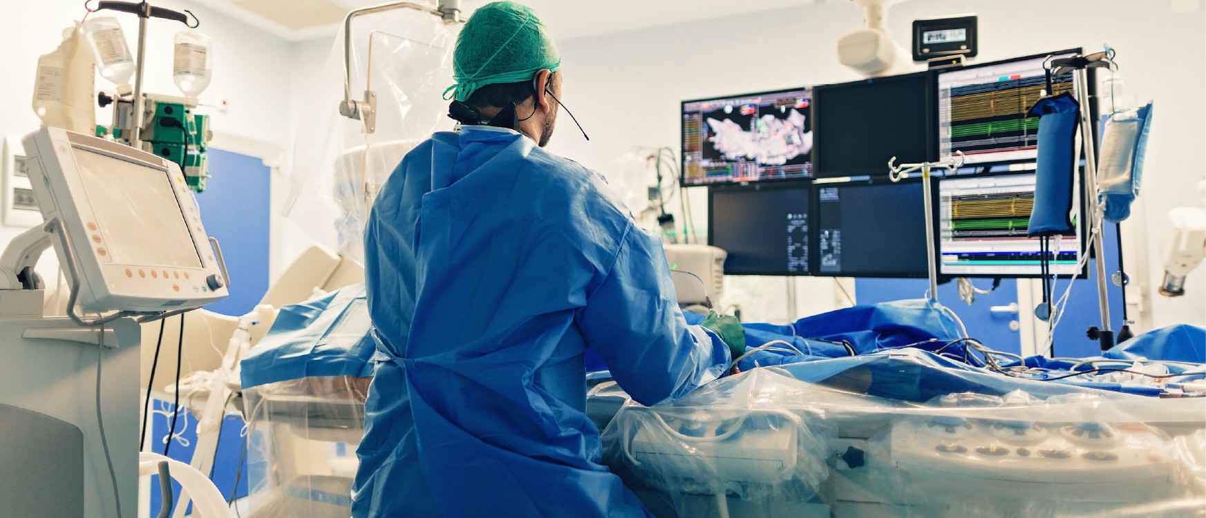 Surgeon in blue scrubs and mask handling a catheter delivery device over a draped patient during an interventional procedure