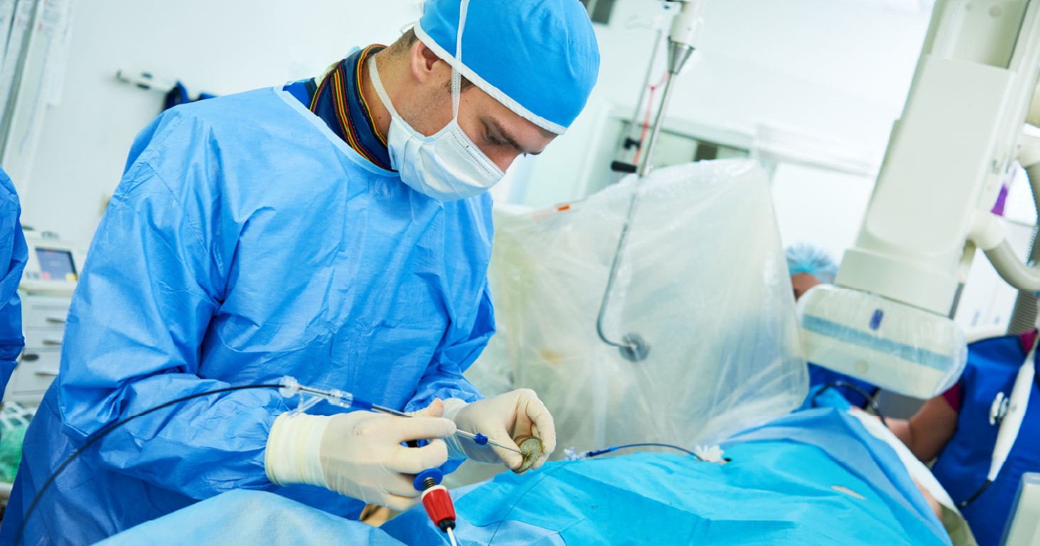 Surgeon in blue scrubs and mask handling a catheter delivery device over a draped patient during an interventional procedure