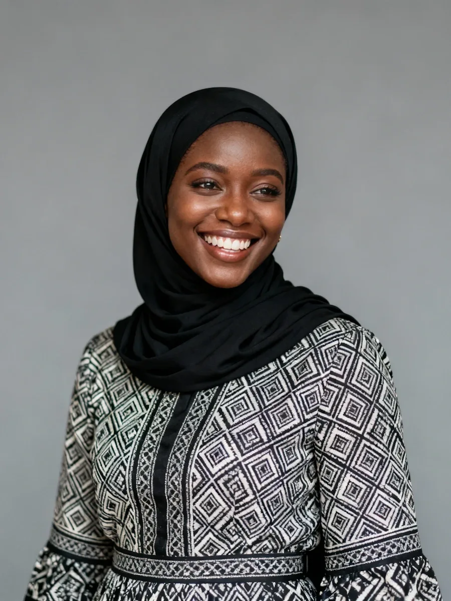 Smiling woman wearing a black hijab and a patterned black and white dress against a gray background.