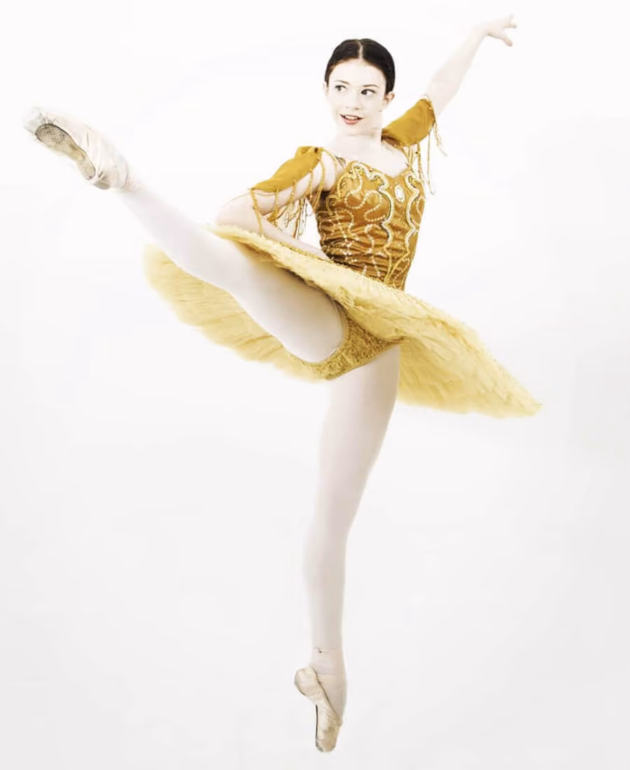 Female ballet dancer in a gold tutu performing an arabesque on pointe with one leg high in the air.