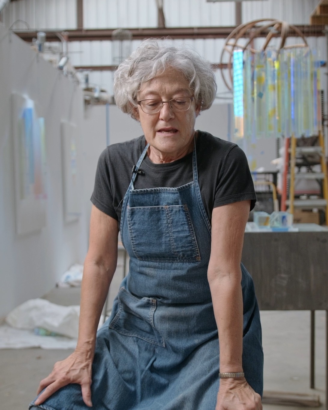 An artist in glasses and a denim apron speaks on camera in her studio, a lavalier mic clipped to her shirt and glass artwork hanging behind her.