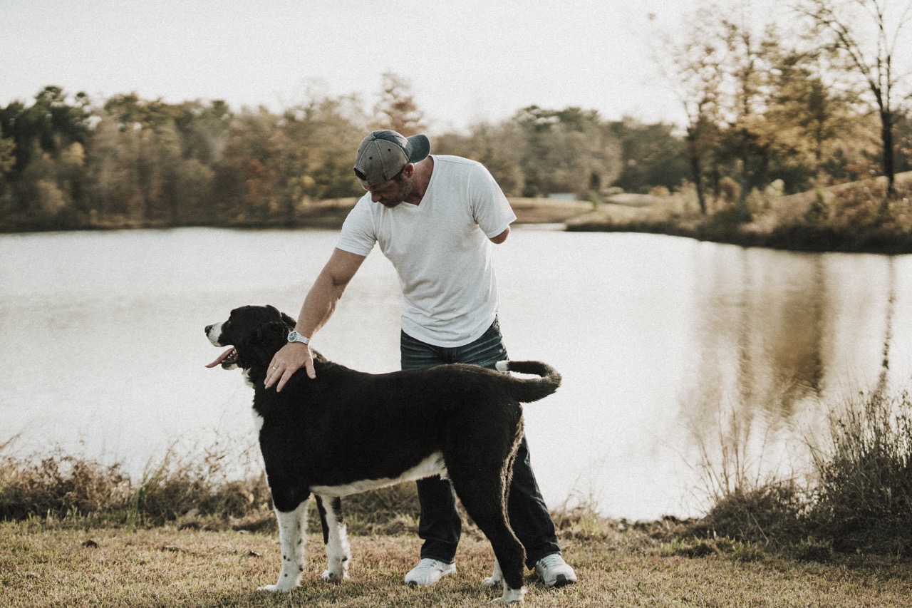 A man with an amputated arm wearing a baseball cap and t-shirt pets a large black-and-white dog near a calm lake.