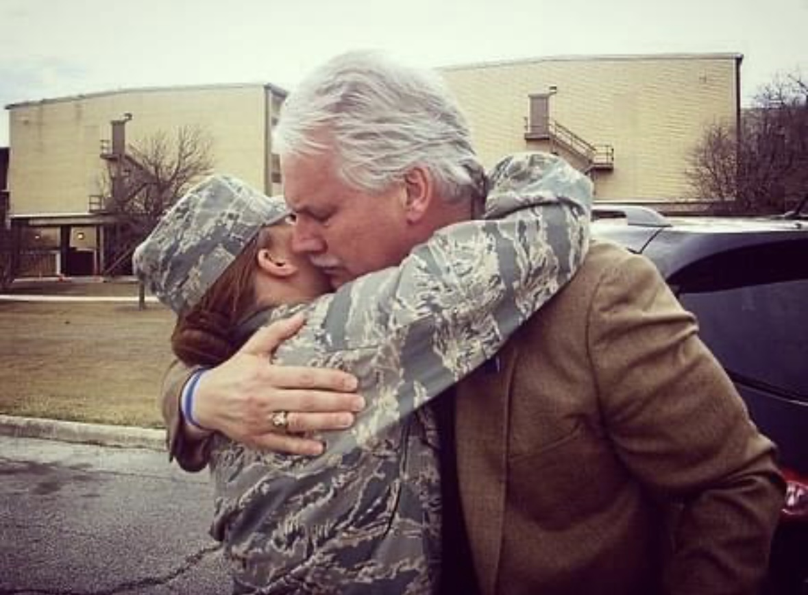 An older man in a jacket warmly embraces a service member in a camouflage uniform and cap outdoors.