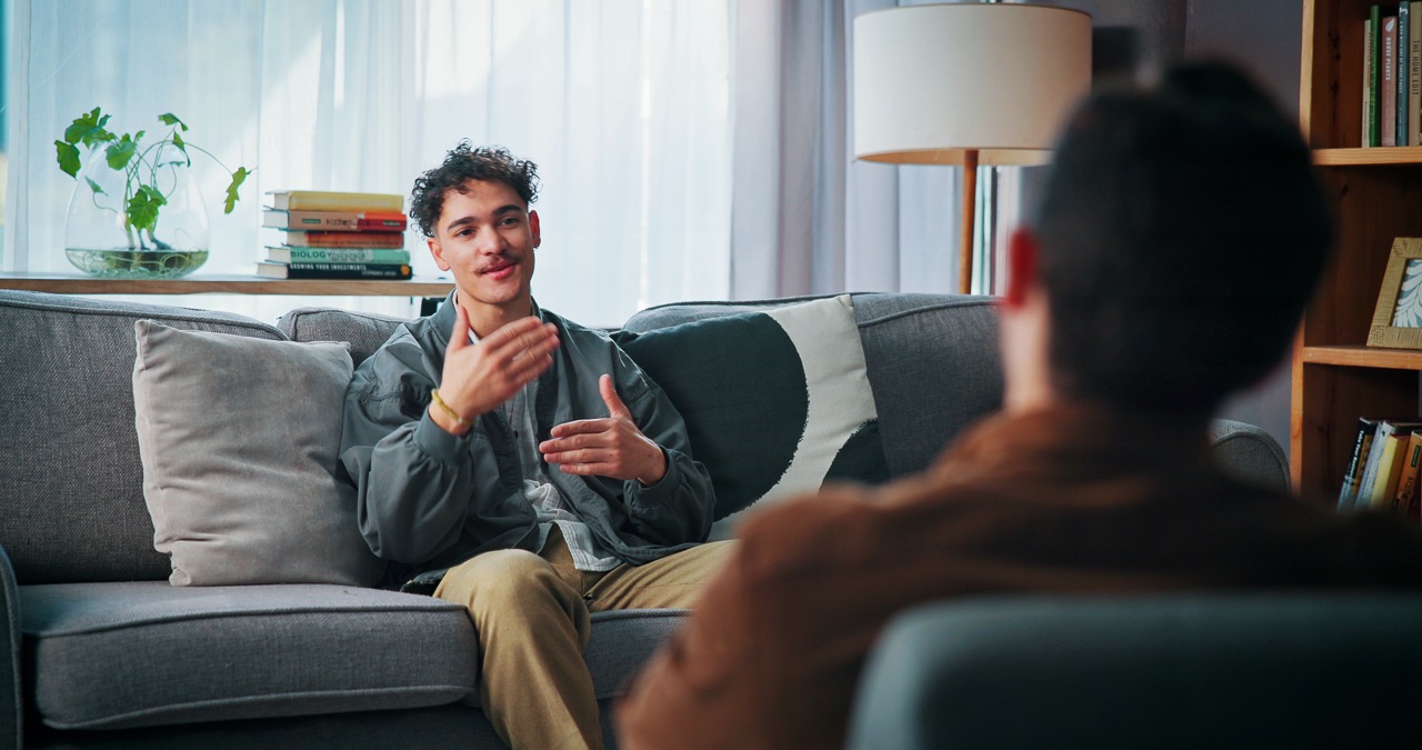 A young man sits on a couch gesturing while speaking to a therapist during an in-person session.