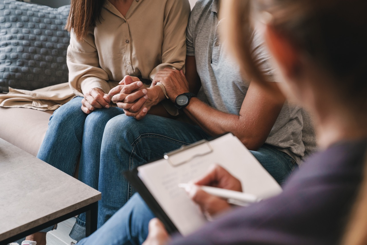 A couple holding hands on a couch while sitting across from a therapist taking notes on a clipboard.