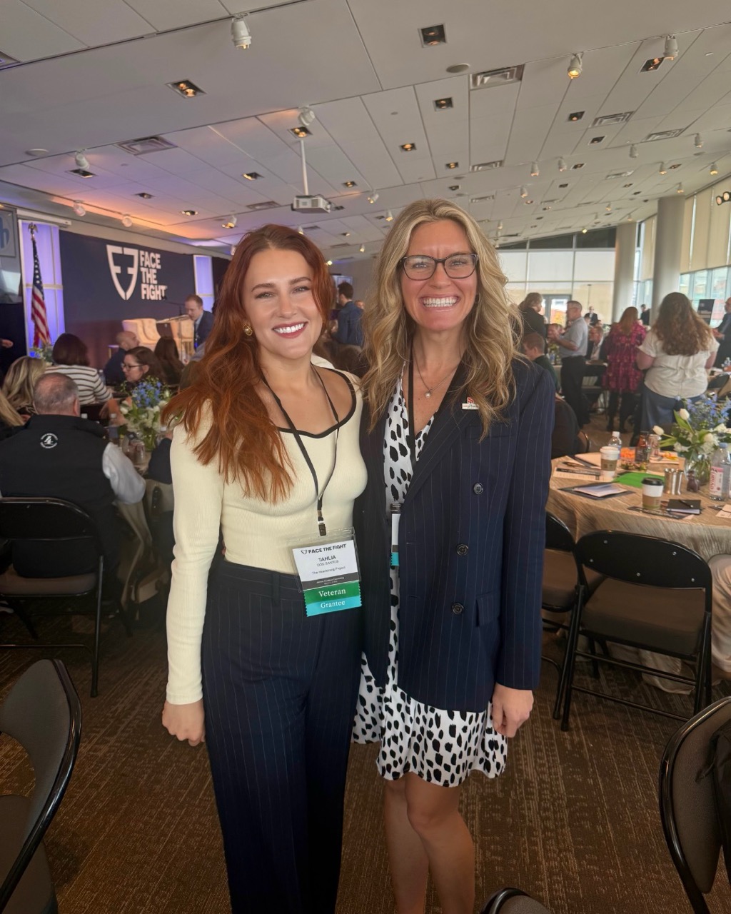 Two women smiling together while attending a conference event inside a large room with tables.