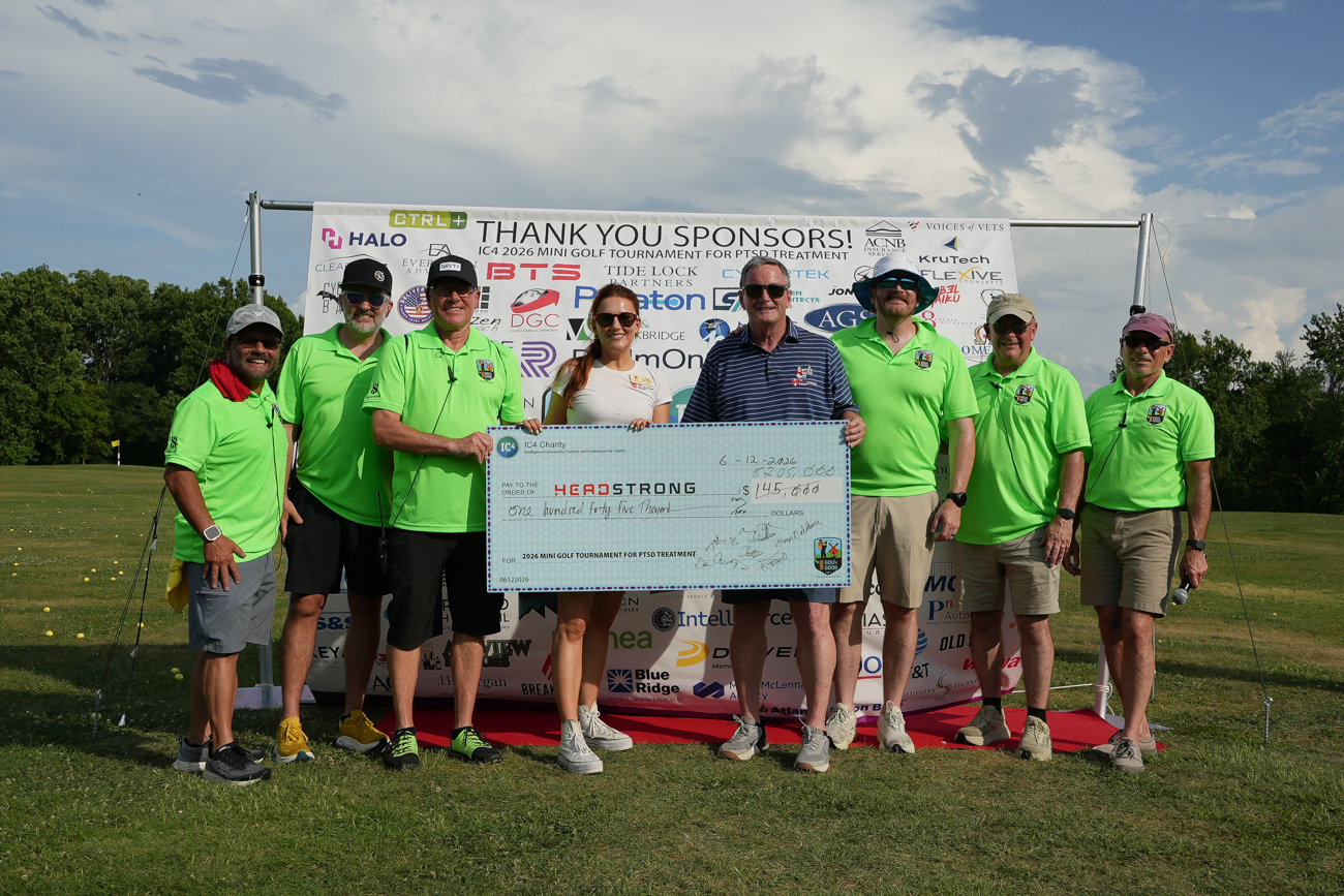 A group of people outdoors holding a large donation check presented to The Headstrong Project in front of a sponsor banner.
