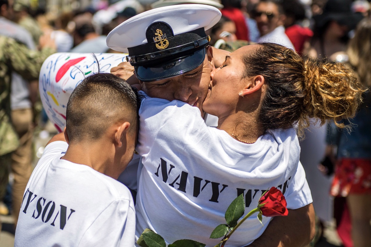 A U.S. Navy sailor in dress uniform smiles while being warmly hugged by his family, as a woman kisses his cheek.