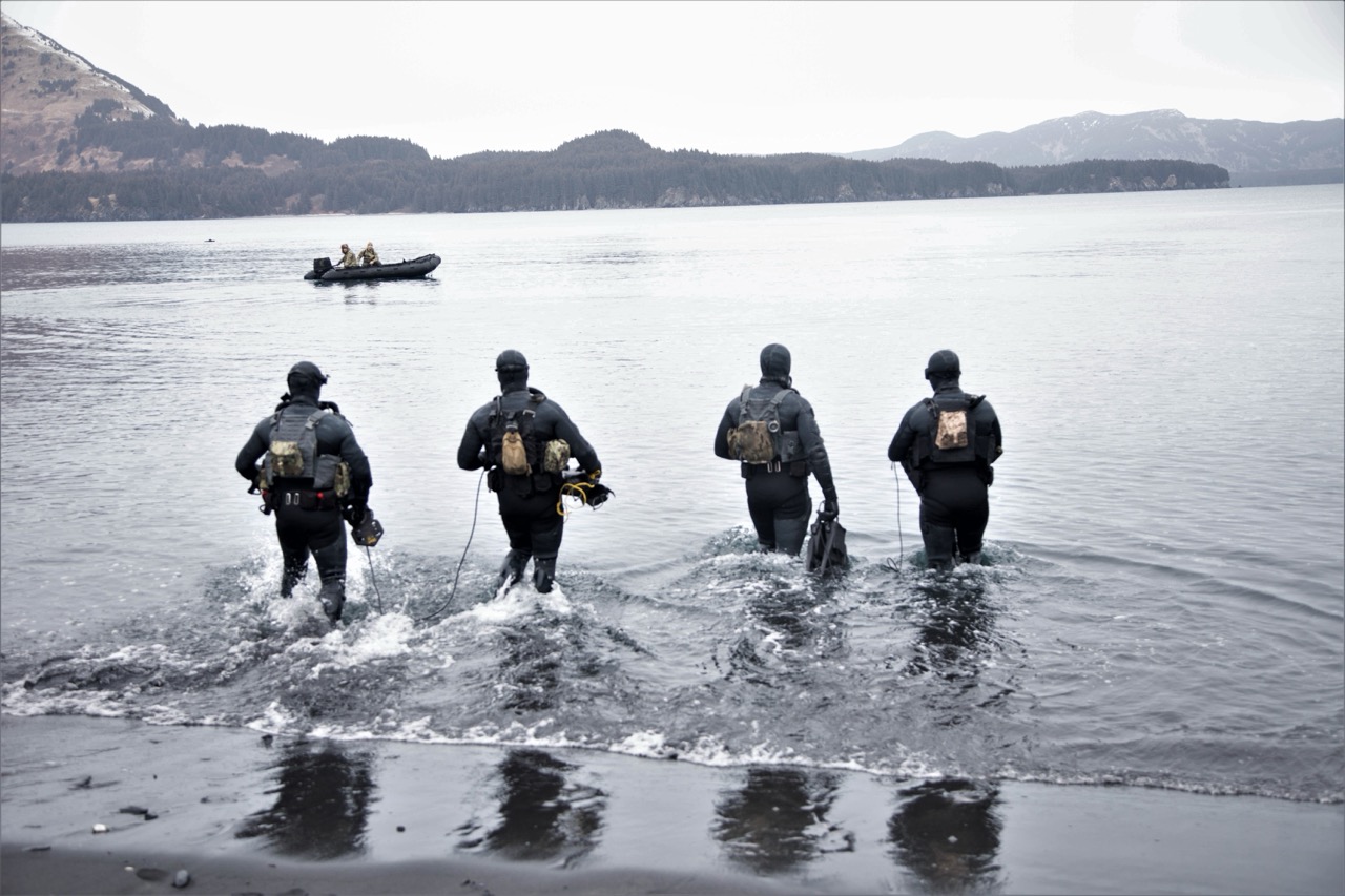 Four military divers in wetsuits and tactical gear wade into cold open water from a shore toward a small boat.