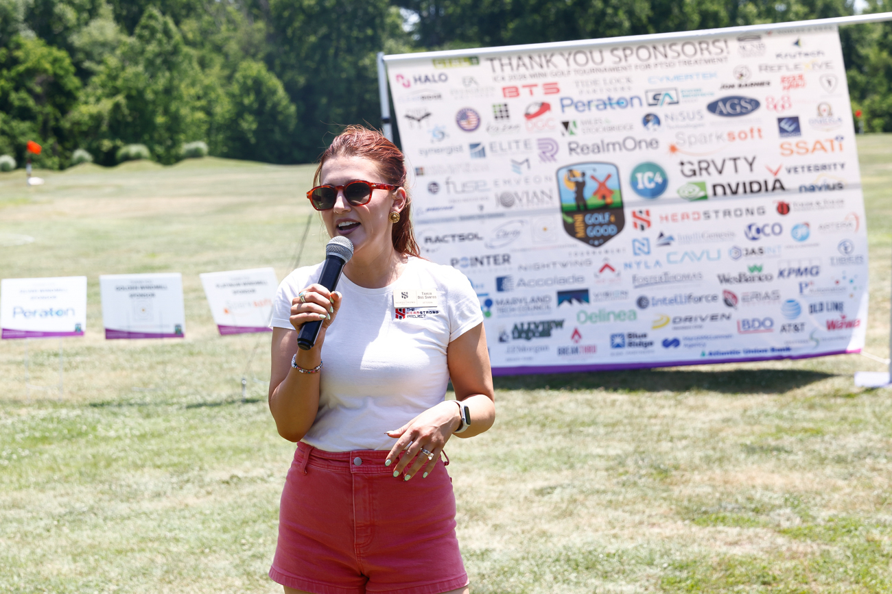 A woman speaks into a microphone outdoors in front of a sponsor banner at a charity event.