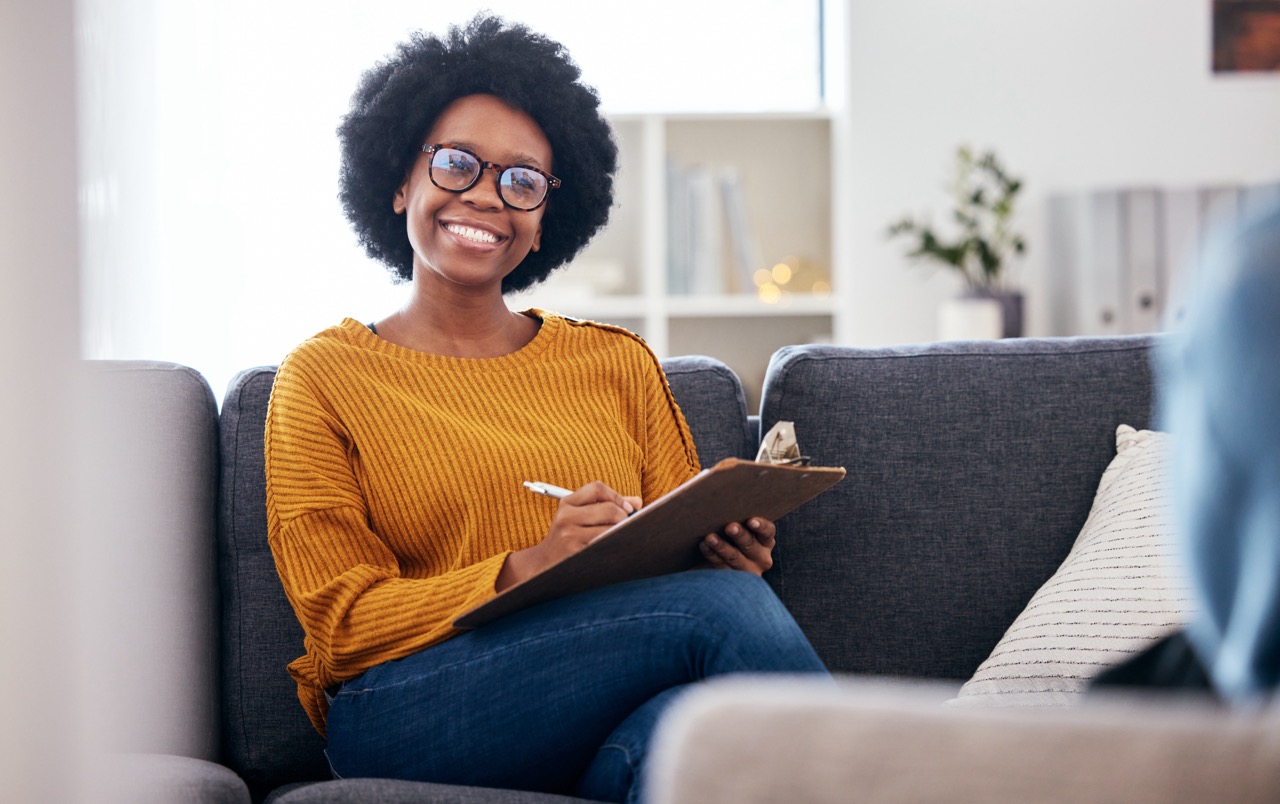 A smiling female therapist wearing glasses sits on a couch while holding a clipboard and pen.