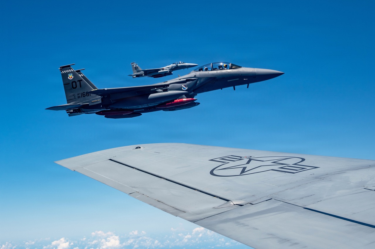 Two U.S. military fighter jets flying in formation against a clear blue sky, viewed over the wing of another aircraft.