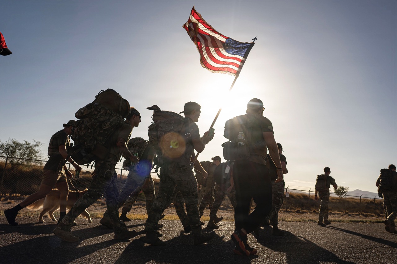 A group of people carrying heavy rucksacks march along a road in silhouette, with one participant holding a waving American flag against the bright sun.