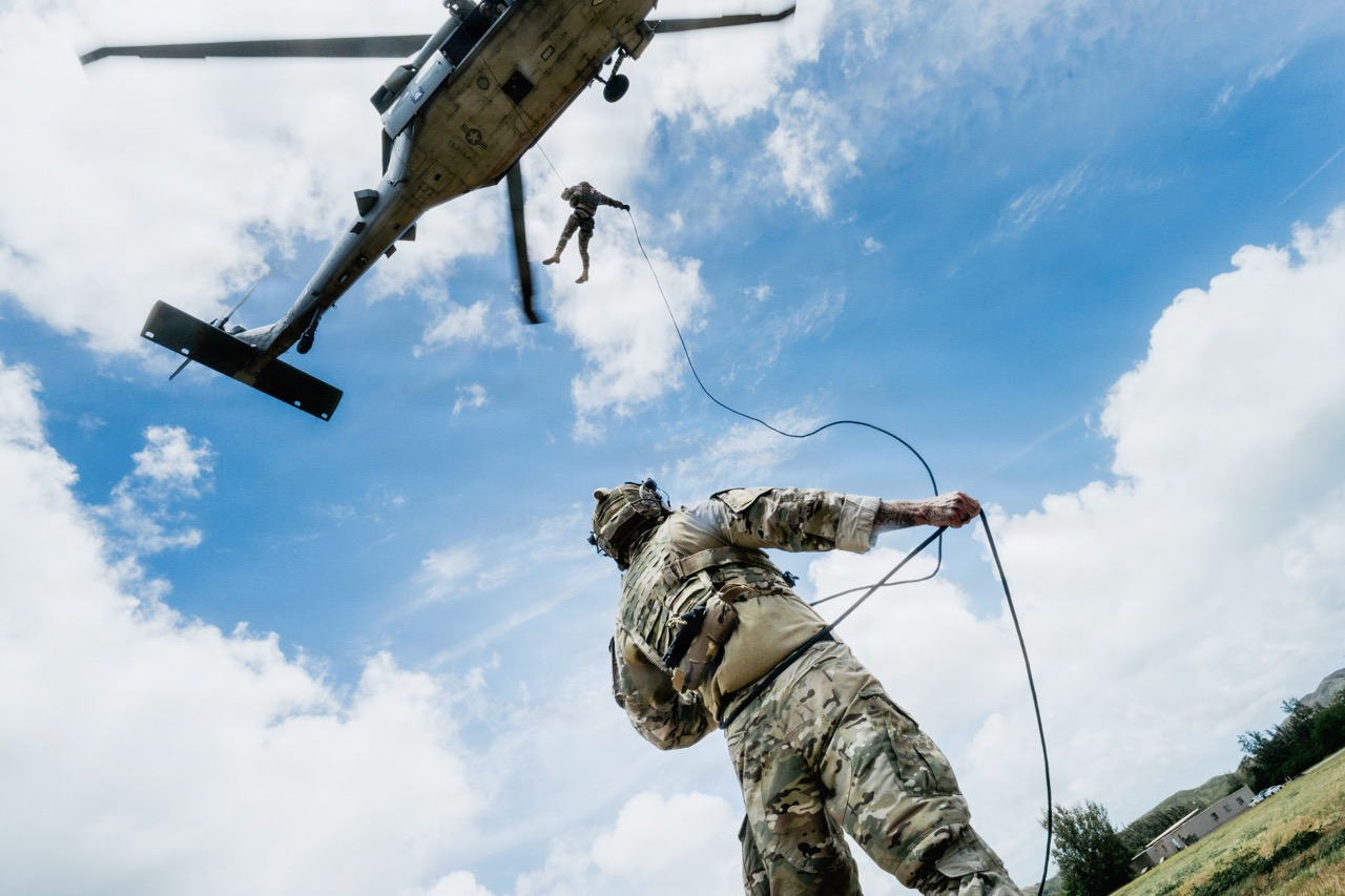 Low-angle view of a military service member holding a rope on the ground as another soldier rappels down from a military helicopter overhead against a partly cloudy blue sky.