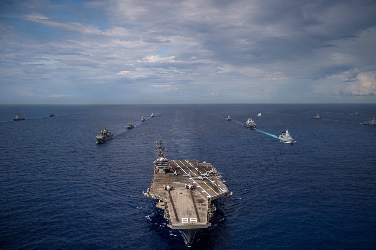 High-angle view of a large naval fleet sailing in formation across open water, led by an aircraft carrier in the foreground under a cloudy sky.
