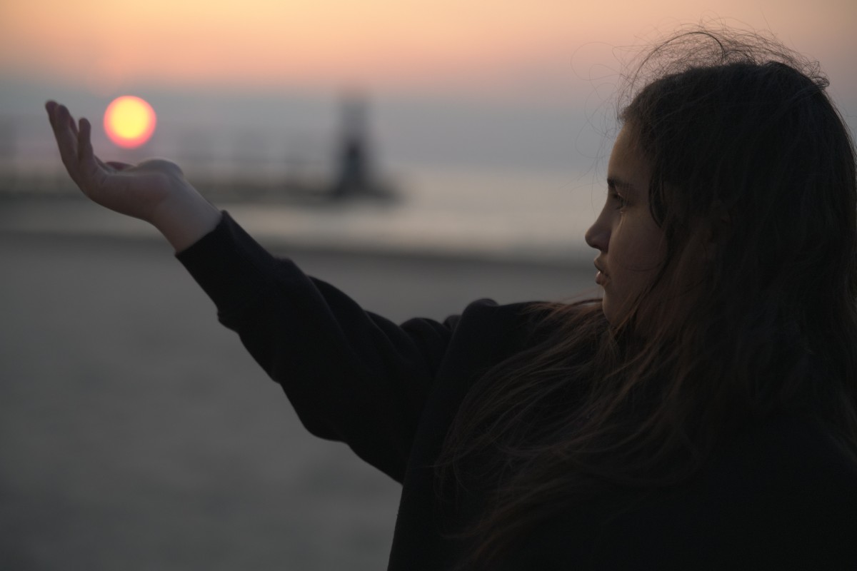 A person holding an outstretched hand beneath the setting sun beside the water.