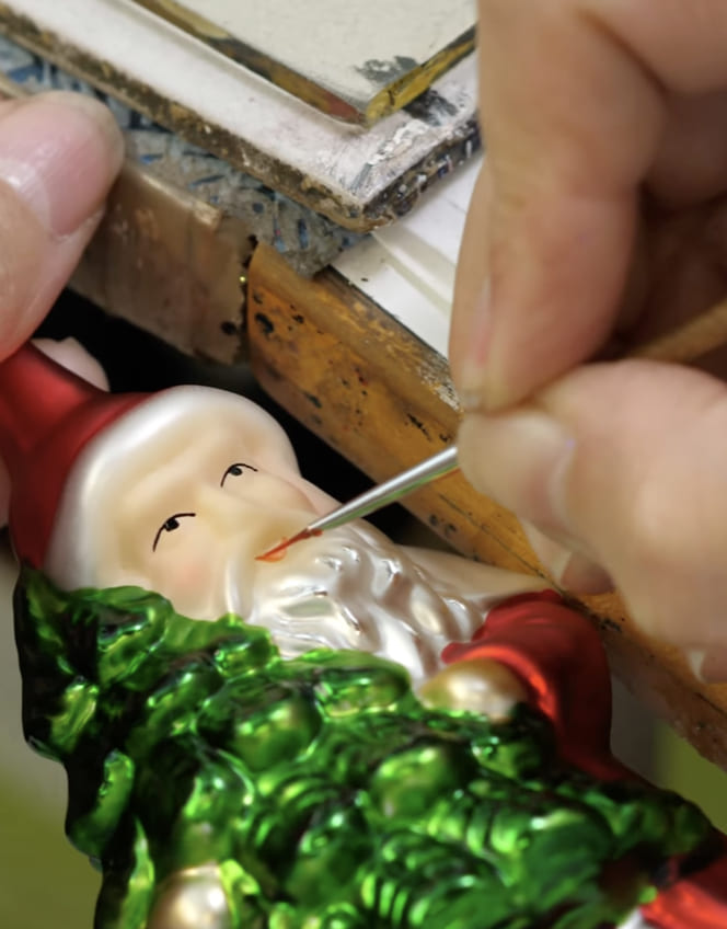 Ein figürliches Glasornament wird mit einem feinen Pinsel bemalt.