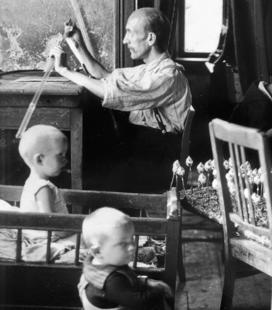 Historische Schwarzweißaufnahme einer Frau bei der Glasbearbeitung mit Kindern an der Werkbank.