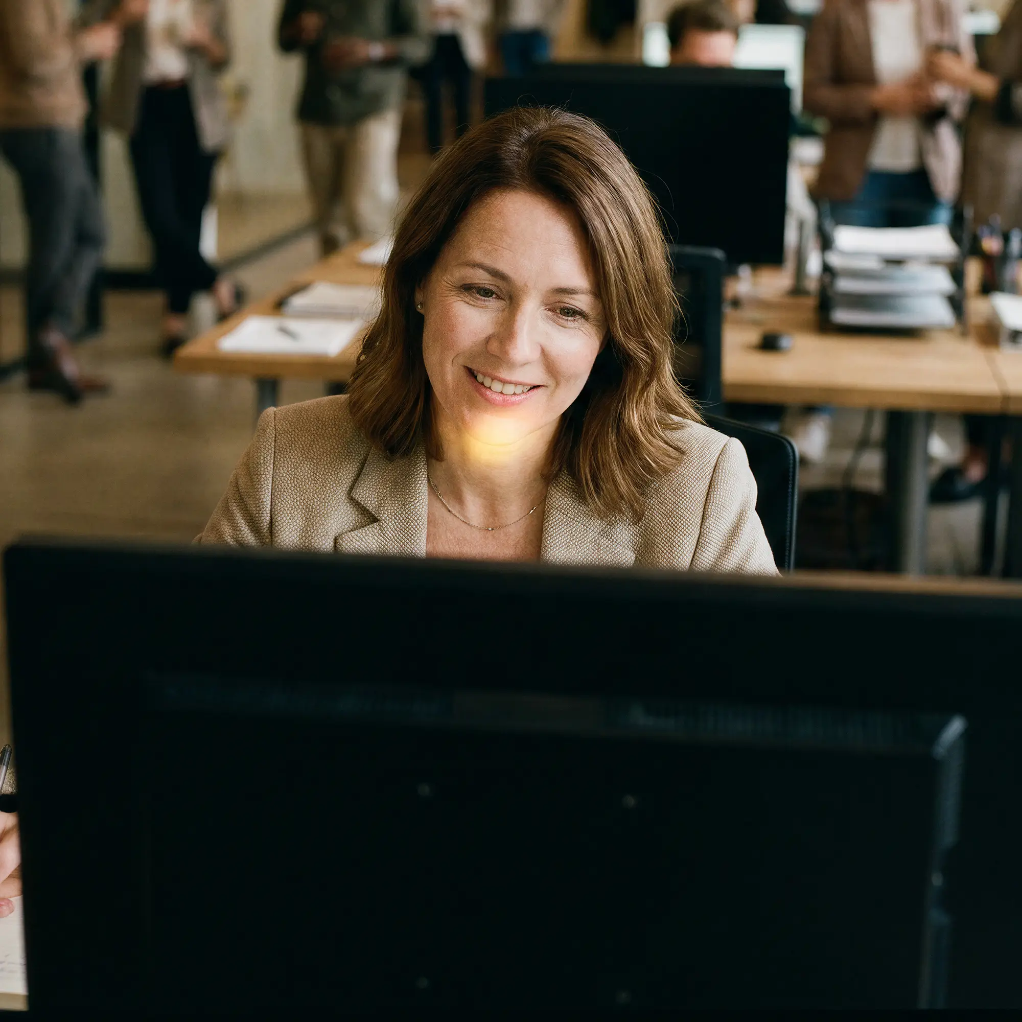 Smiling woman with brown hair working on a computer in a busy office with coworkers in background.
