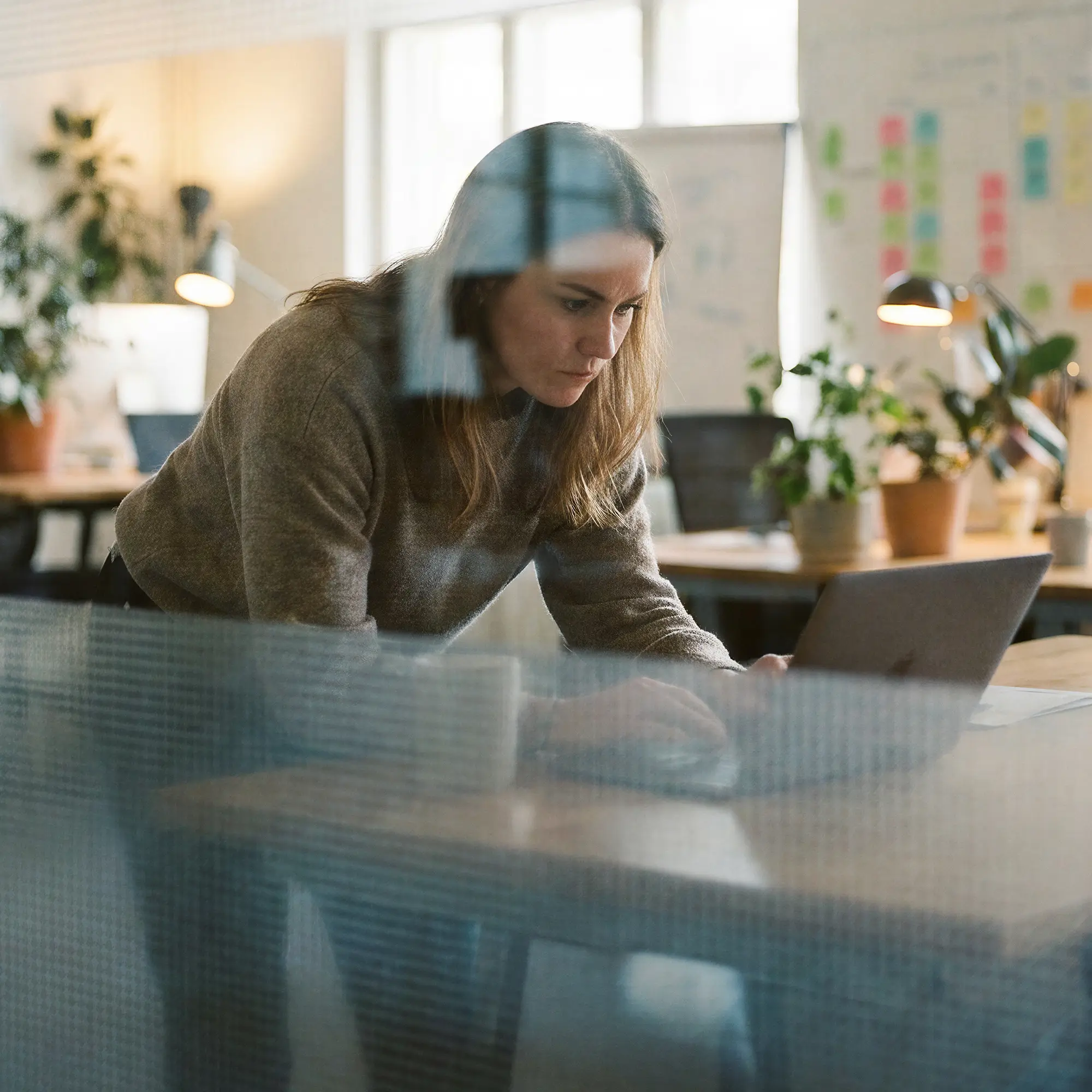 Woman leaning over desk working on a laptop in a bright office with plants and sticky notes.