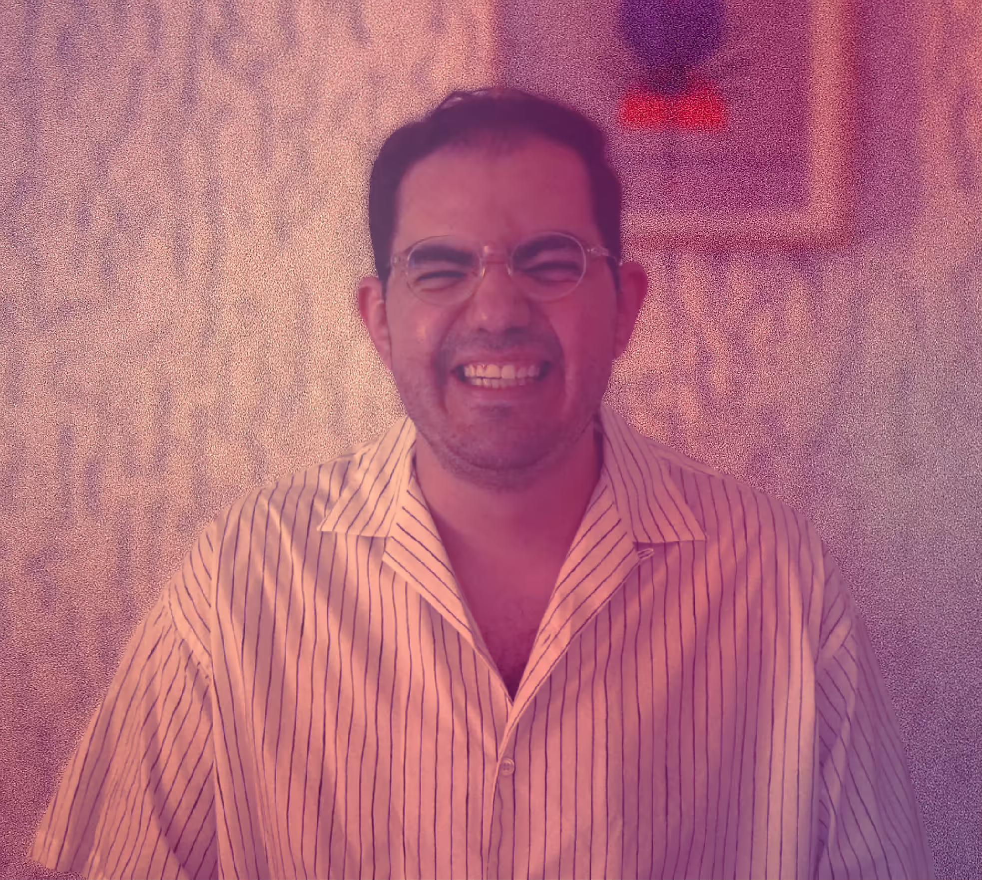 Man with glasses smiling broadly wearing a white striped shirt against textured wall background.