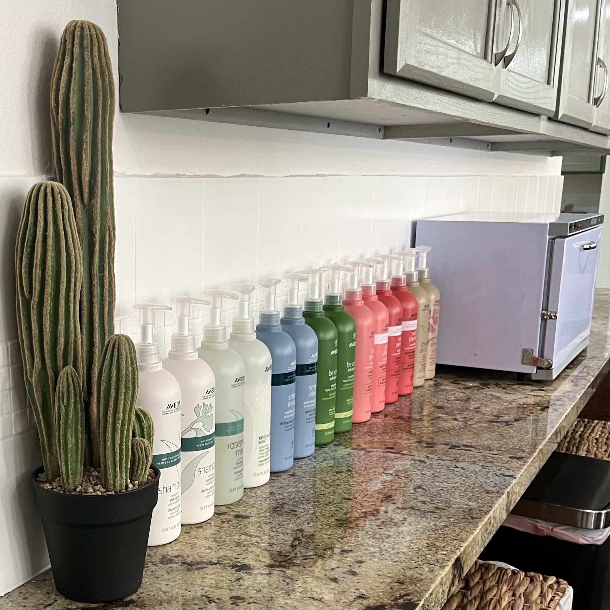 A row of colorful Aveda shampoo and conditioner bottles lined up on a granite countertop next to a black potted cactus and a small lavender-colored machine, with green cabinets above.