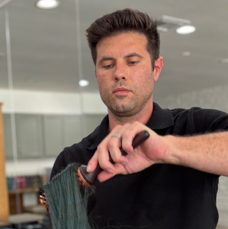 Man with short dark hair wearing a black shirt using a round brush to style wet hair in a modern salon setting.