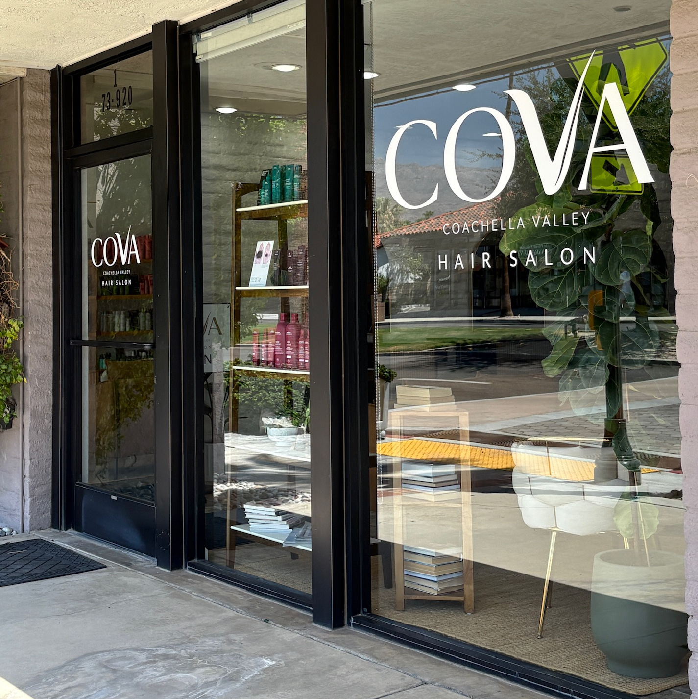Exterior view of COVA Coachella Valley Hair Salon with large glass windows and door, displaying hair care products inside and a tall potted plant by the window.