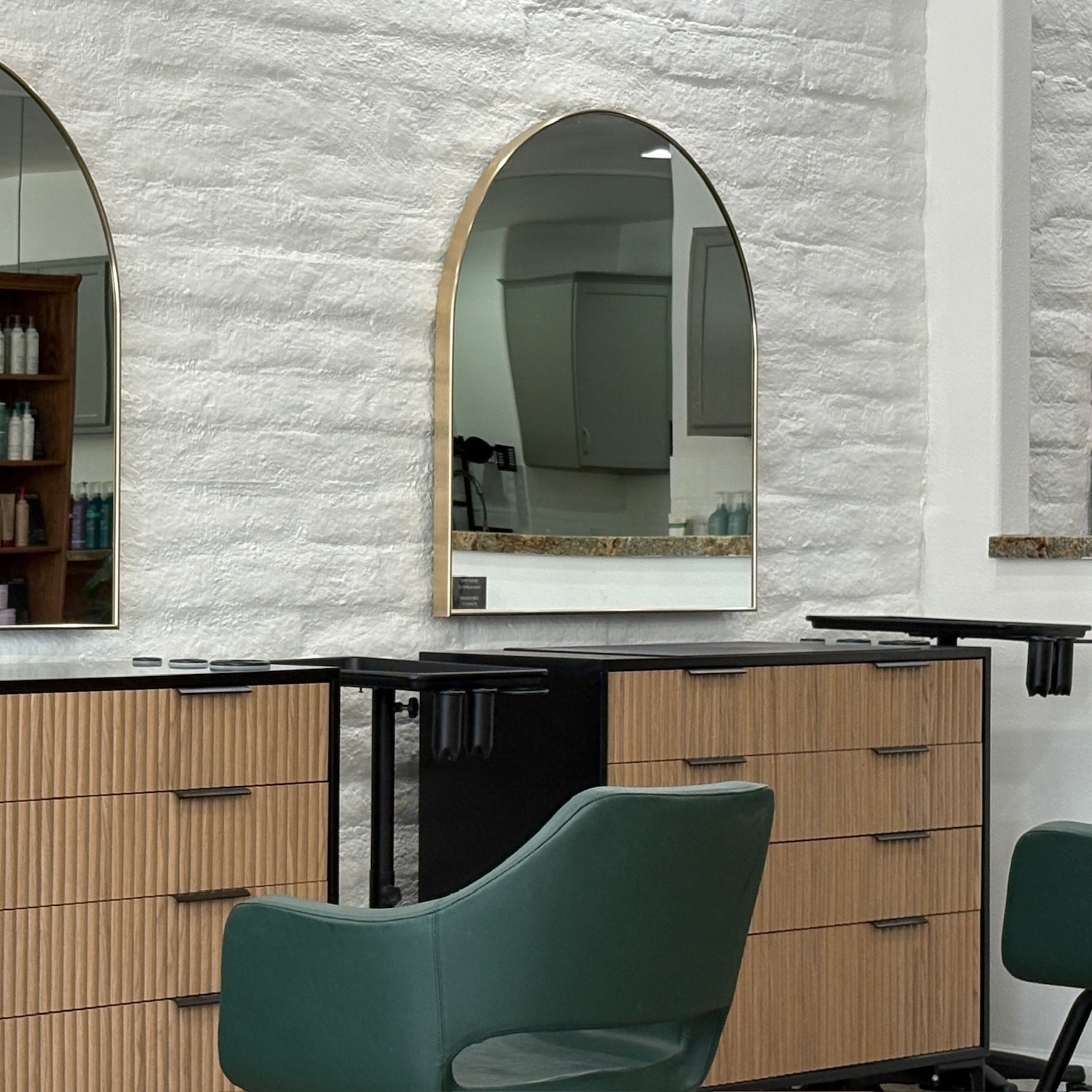 Modern salon interior with green upholstered styling chairs, wooden drawer cabinets with ribbed texture, black metal frames, and arched wall mirrors on a white textured wall.
