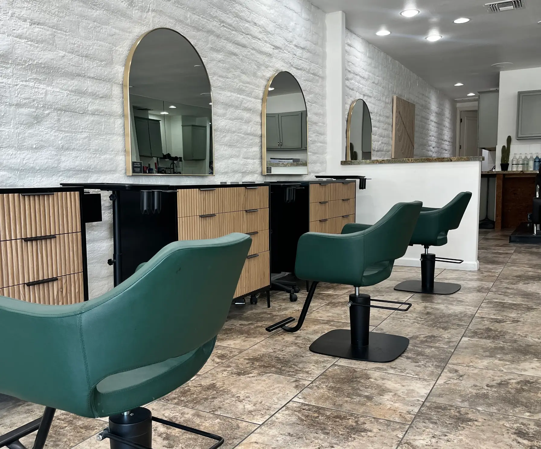 Modern salon interior with three green styling chairs facing large arched mirrors mounted on a white textured brick wall above light wooden cabinets with black handles, on a tiled floor.