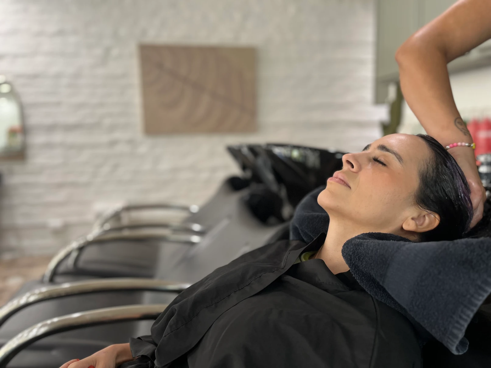 Woman with closed eyes reclining at a salon sink as a stylist washes her hair with black towels around her neck.