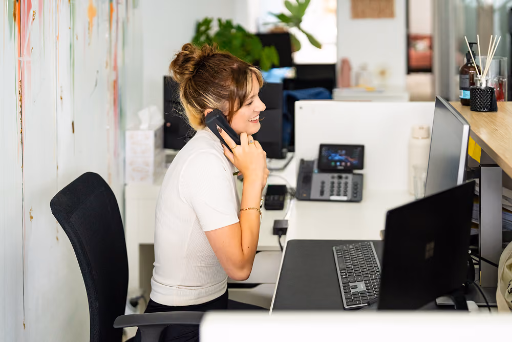 Woman smiling while talking on the phone at her desk with a computer and office phone.