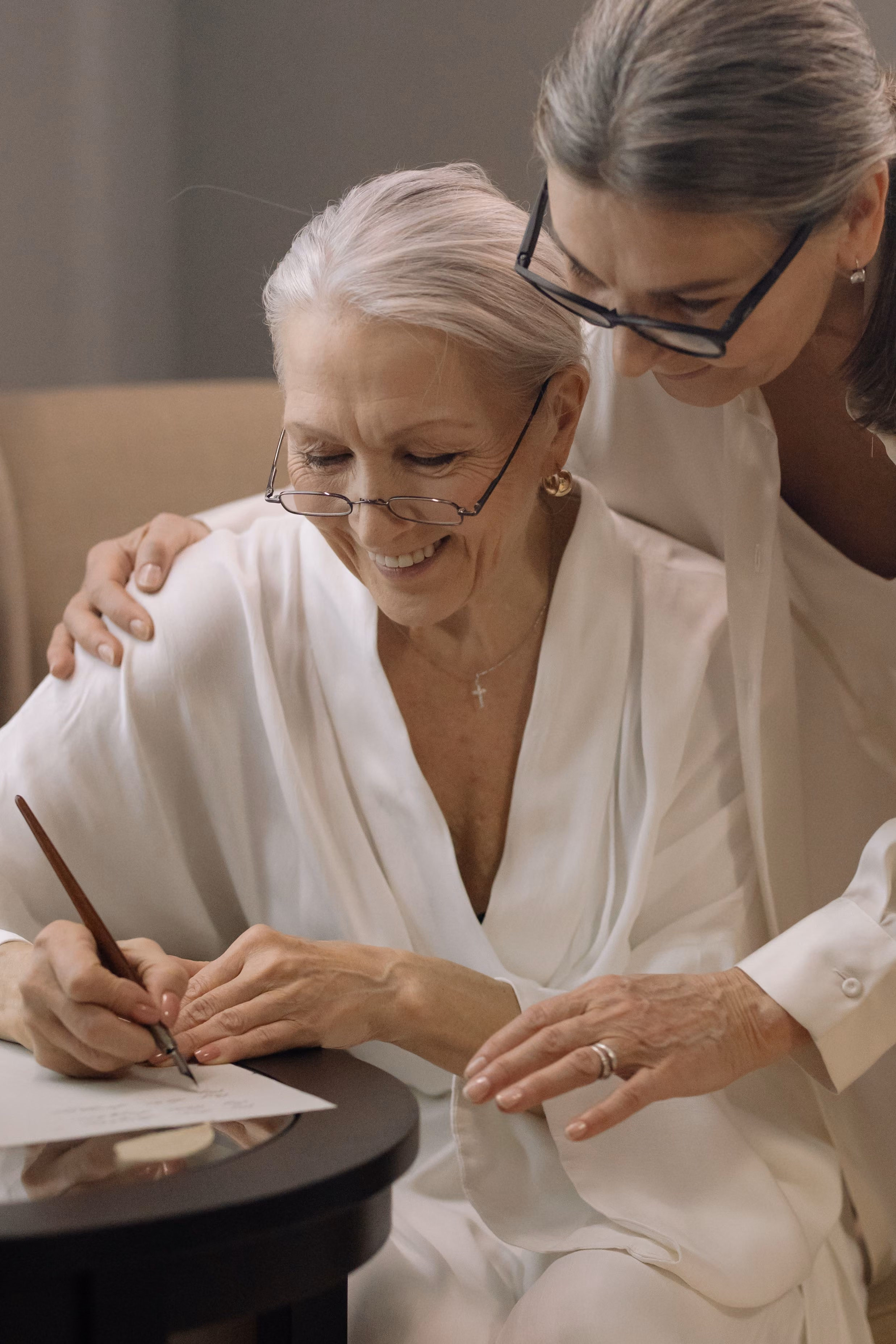 Two older women in white are smiling, one is seated signing a Power of Attorney document while the other leans over her.