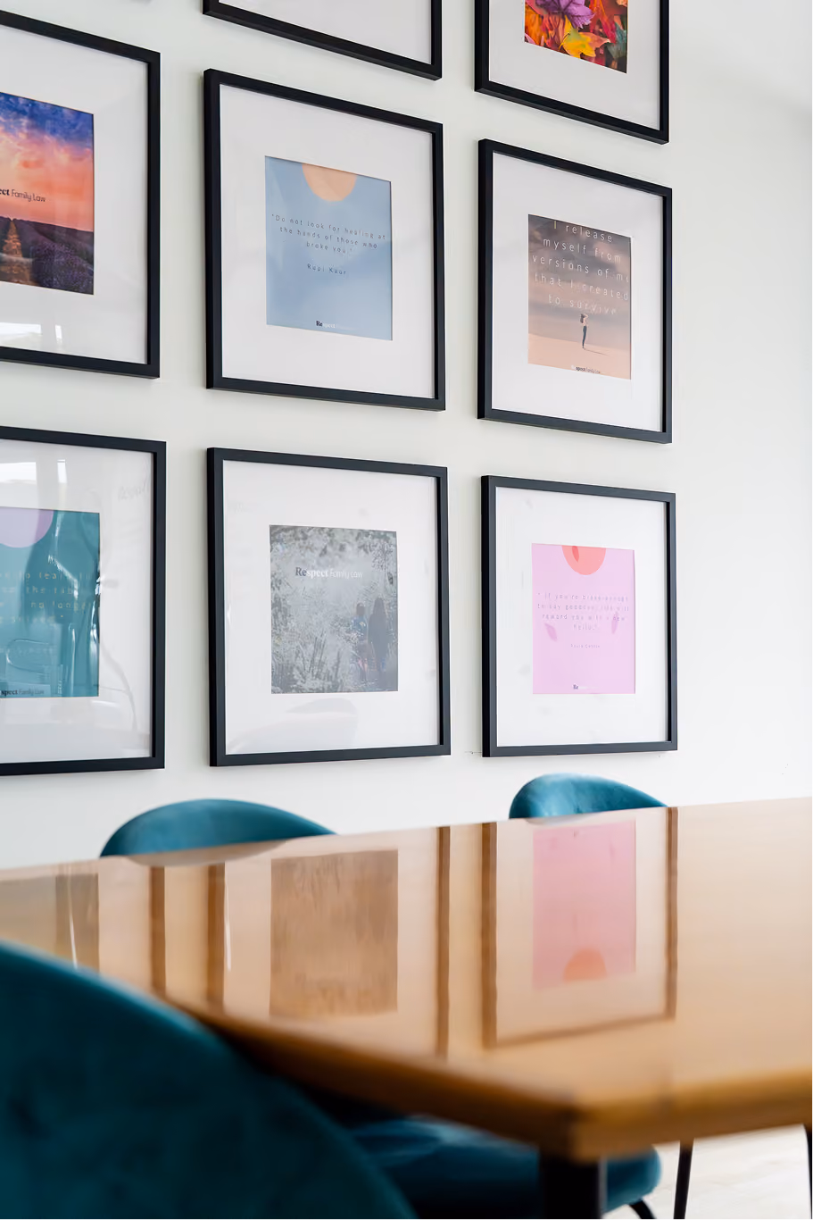 Wooden table and teal chairs in front of a wall with black framed inspirational quotes and art.