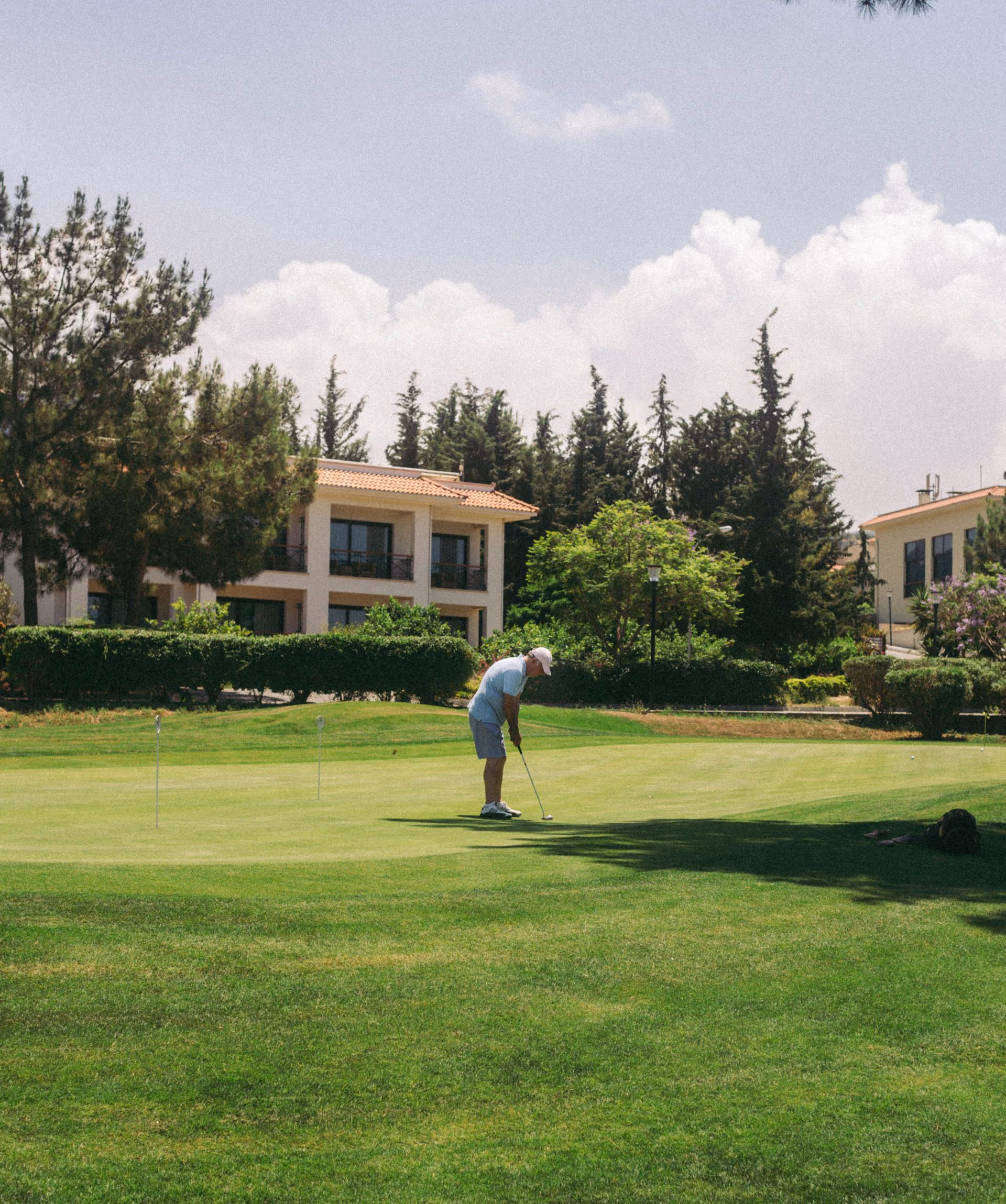 Man in light clothes putting on a green golf course with trees and buildings in background.