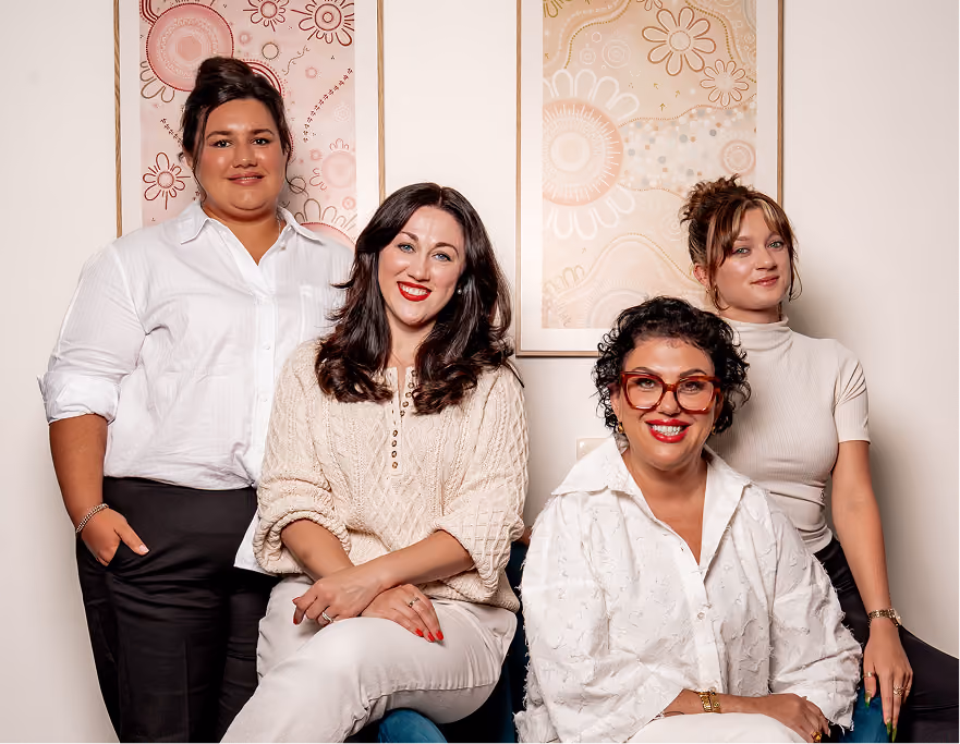 Four diverse women smiling, posing in front of wall art with floral designs, wearing smart casual clothes.