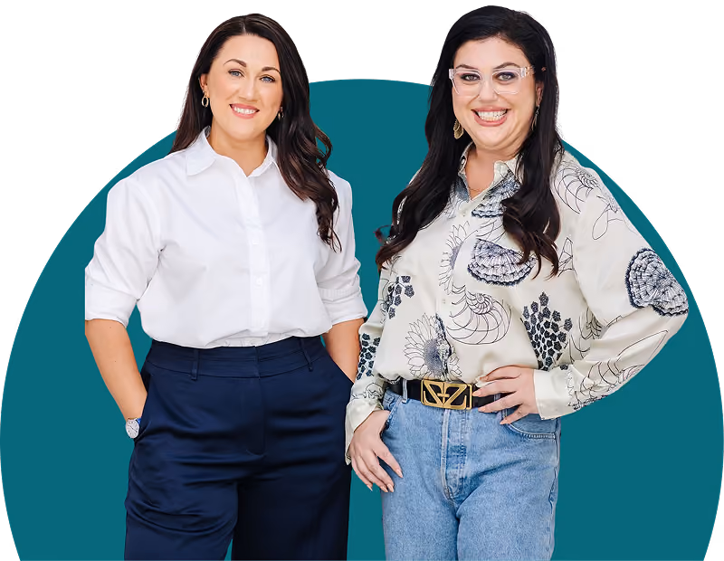 Two smiling women standing, one in white shirt and navy pants, the other in patterned shirt and jeans.