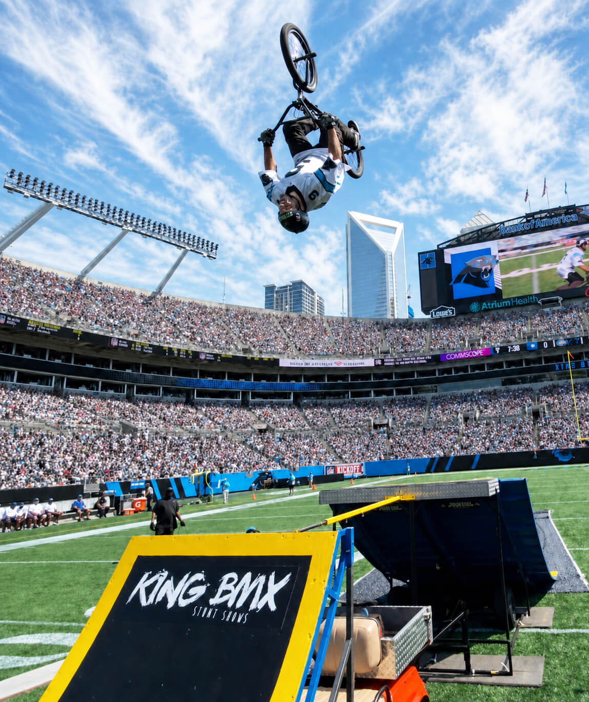 BMX rider in mid-air flip above stadium ramp with a crowd and city buildings in background.