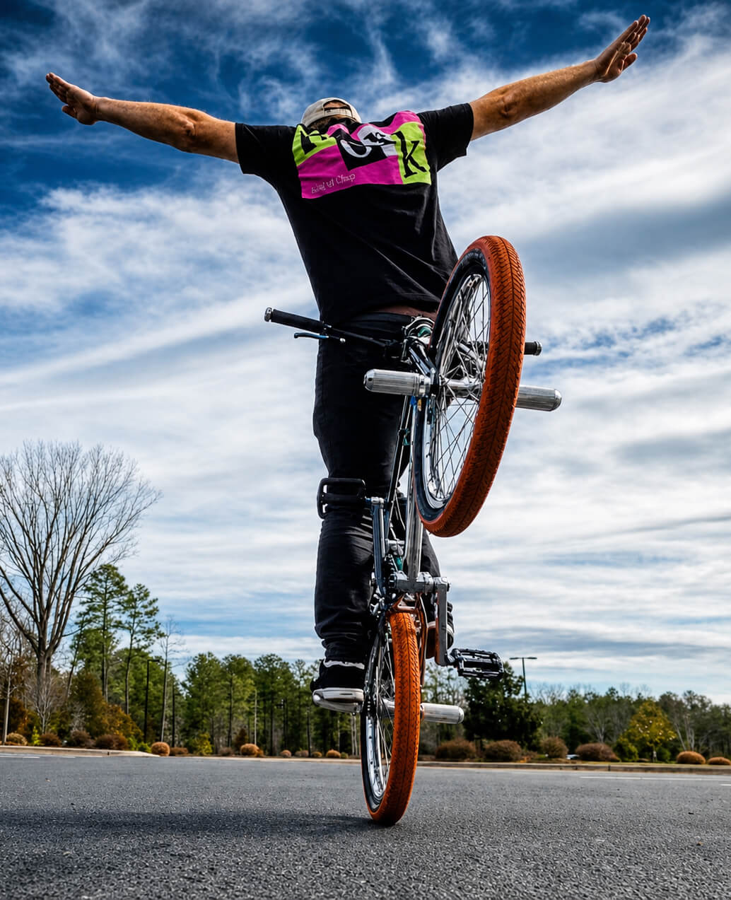Keith King riding a bicycle on one wheel with arms outstretched under a cloudy blue sky.