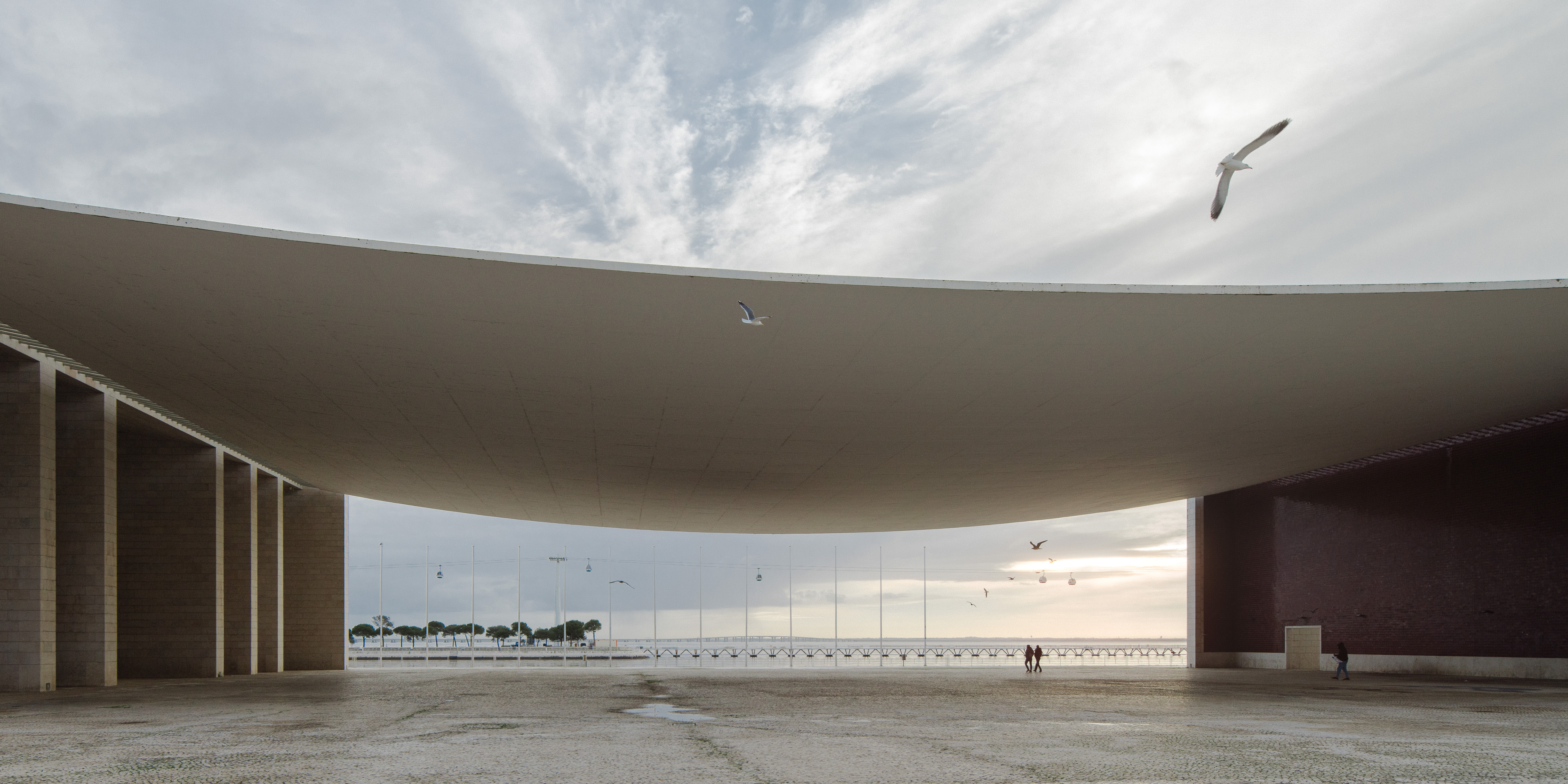 Large curved architectural canopy with seagulls flying and people walking near waterfront at sunset.