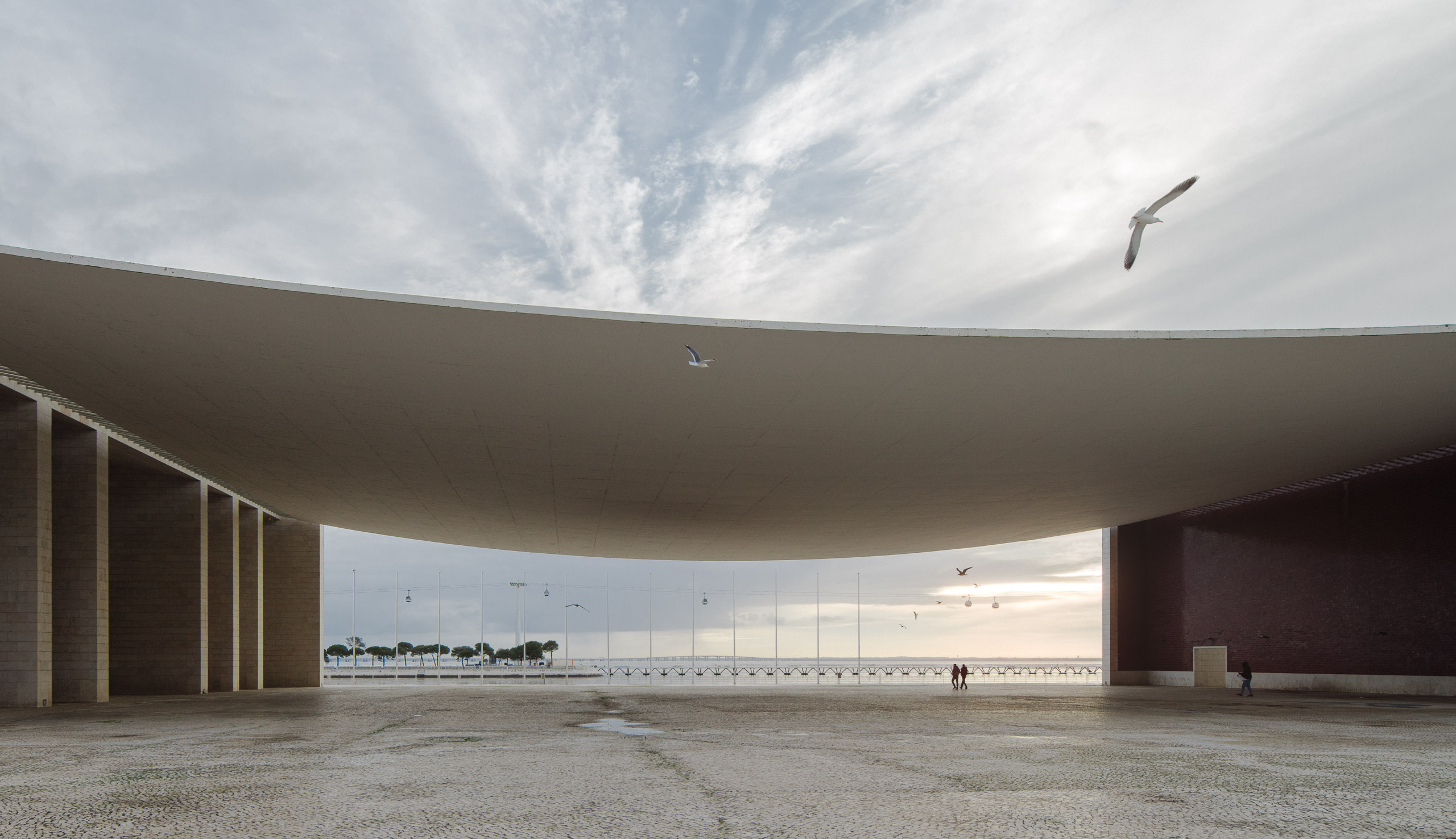 Large curved architectural canopy with seagulls flying and people walking near waterfront at sunset.
