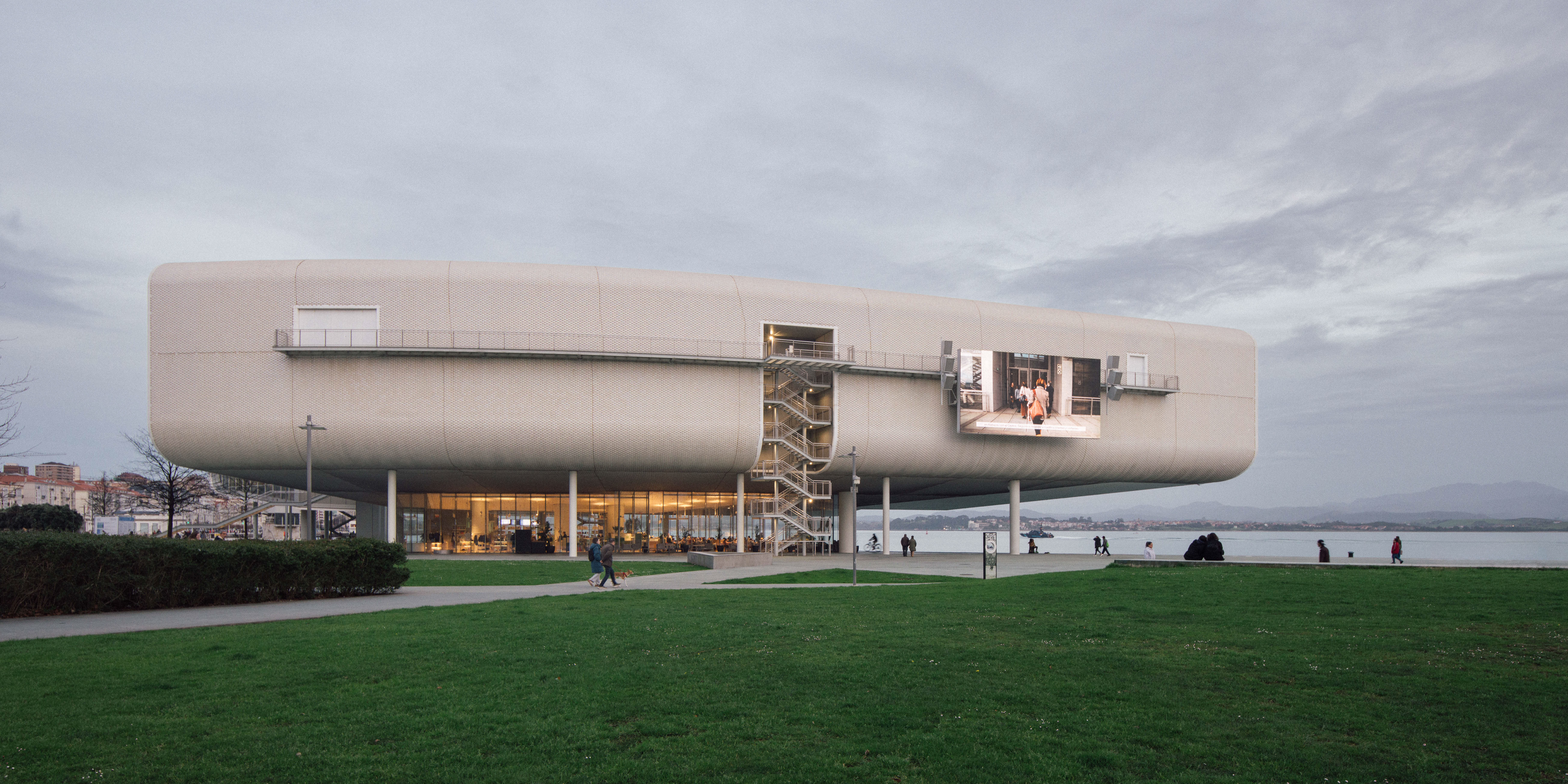 Modern rounded building elevated on pillars beside water with green lawn and people walking nearby.