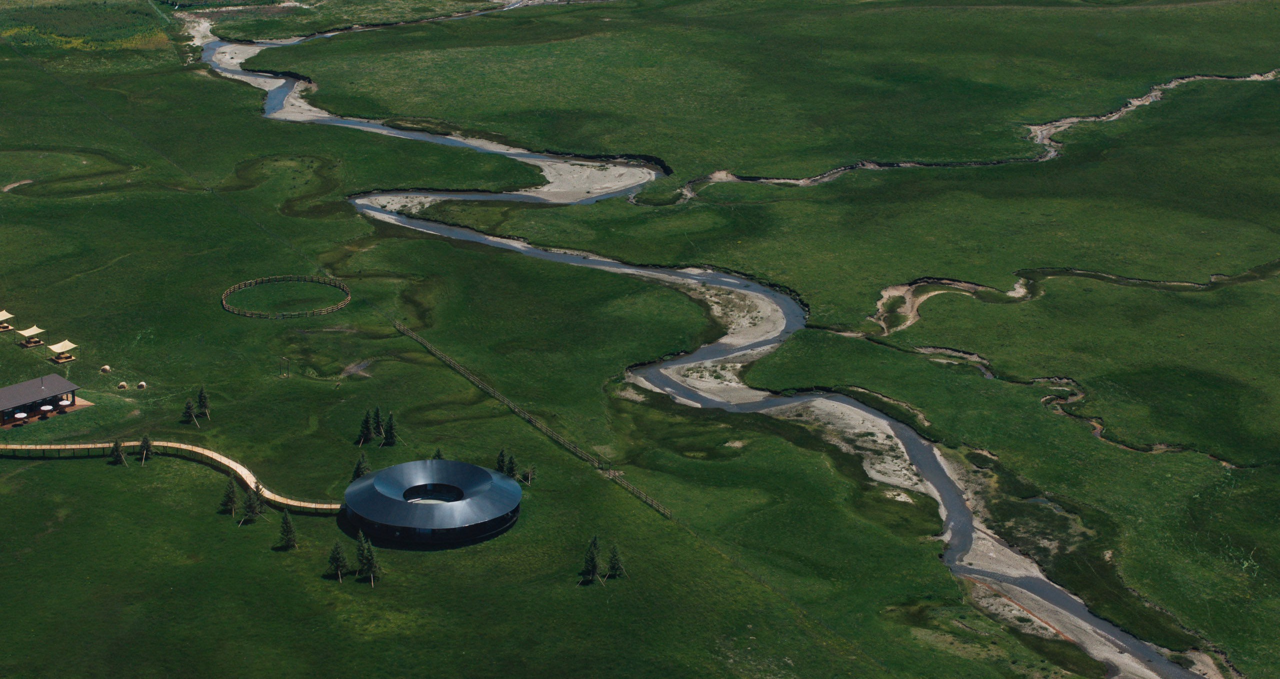 Aerial view of green fields with a winding river, conical trees, and a modern circular building.