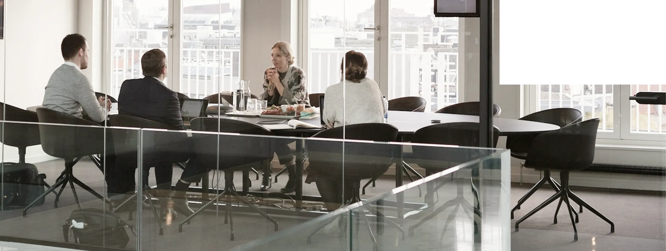 Four people sit around a conference table in a glass-walled meeting room with large windows and office chairs.