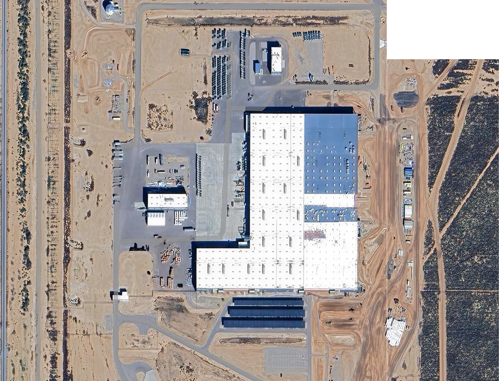 An aerial view of a large industrial building complex with white and blue roofs, surrounded by paved areas, dirt roads, and desert terrain.
