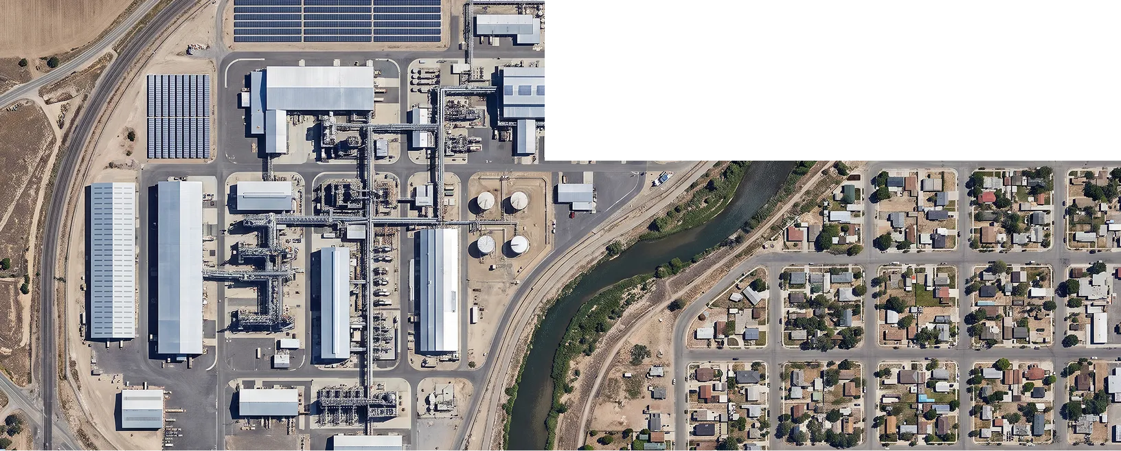 An aerial view of an industrial facility with large buildings, tanks, and solar panels beside a river, next to a grid of suburban homes.